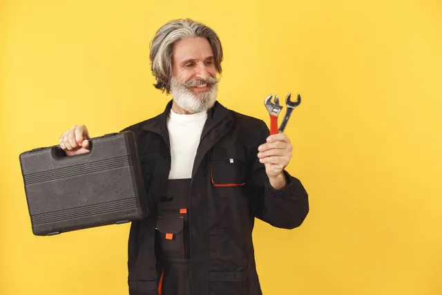 Elderly bearded man with safety glasses smiling, holding a toolbox and wrenches against a yellow background. How Alberta trades licensing affects appliance repair bookings in Calgary and Edmonton